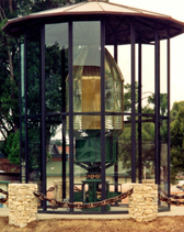 Glass enclosure surrounding a lens with conical top, curved glass panels, and clockwork base.