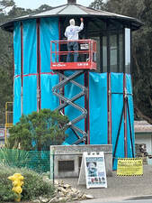 A conical glass enclosure is covered in protective blue sheeting, while a man on top of a scissorjack works on the enclosure.