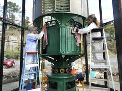 A conical green lens and mechanism is wiped by two volunteers on ladders, with the glass enclosure surrounding them.